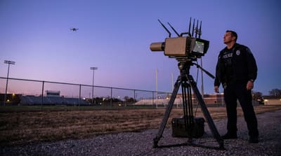 A police-operated counter-drone detection unit on a tripod scanning the sky near a fenced stadium at dusk, with a small commercial quadcopter visible in the distance holding position