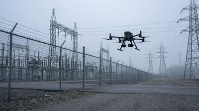 Commercial inspection drone holding position at a distance from a fenced electrical substation and industrial facility under an overcast morning sky, with transmission towers receding into the background