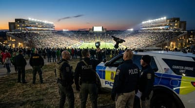 Law enforcement counter-drone team monitoring airspace near a packed stadium at dusk, handheld detection equipment raised toward the sky