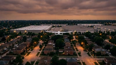 A small package delivery drone in wing-borne flight above a Chicago-area suburban neighborhood at dusk, warehouse rooftops and residential streets receding under an overcast sky